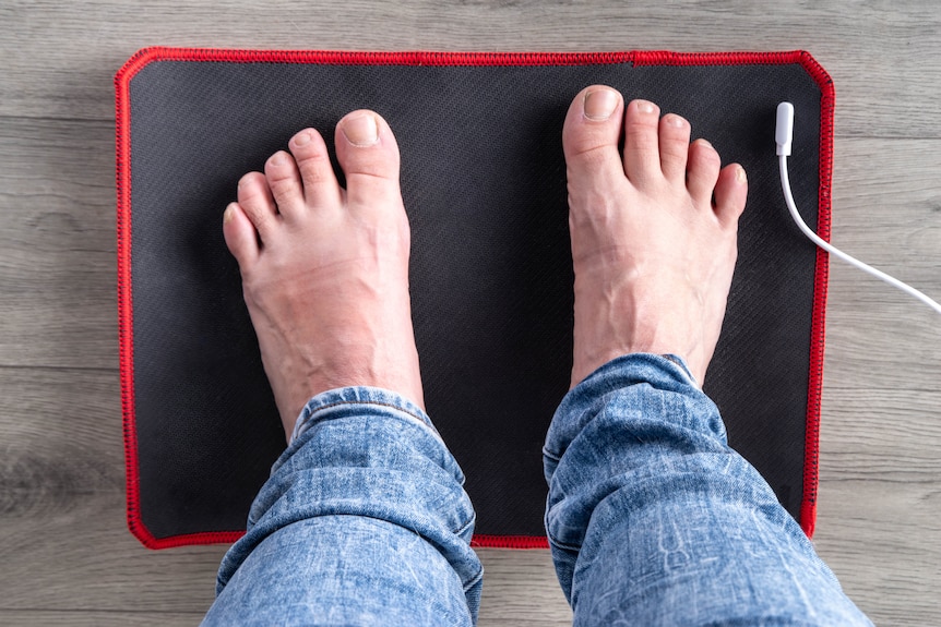 A man in jeans with bare feet on a black mat. There is a white cord attached to it.